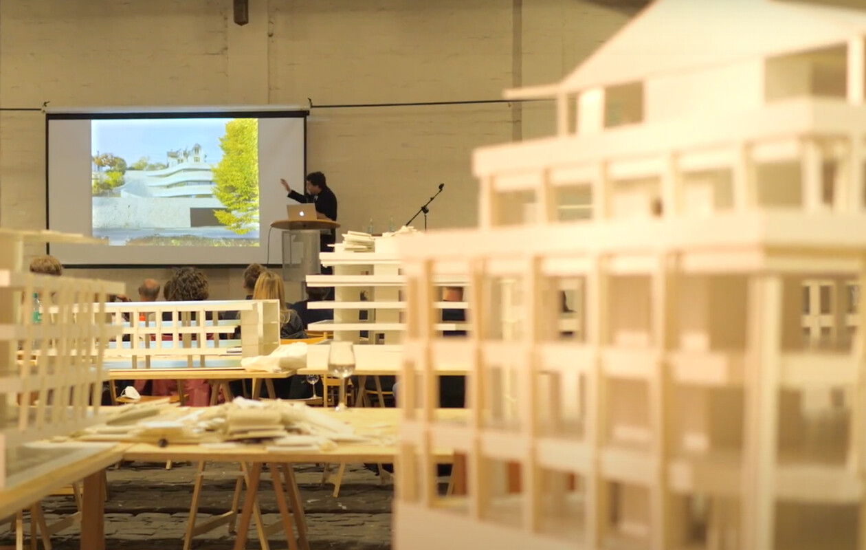Vortragssituation mit Leinwand, im Vordergrund unscharfe Architekturmodelle.