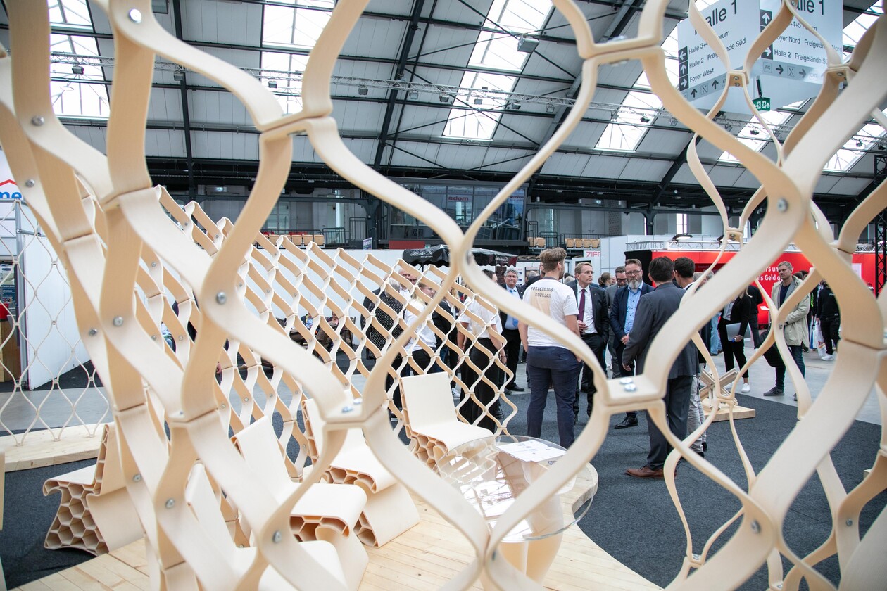 Blick durch eine organische Holzstruktur auf Besucher einer Messehalle.