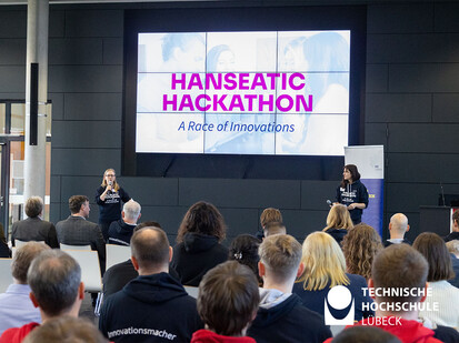 Einleitende Worte in den Hackathon Zwei Personen stehen vor einer Präsentation und das Publikum guckt gespannt