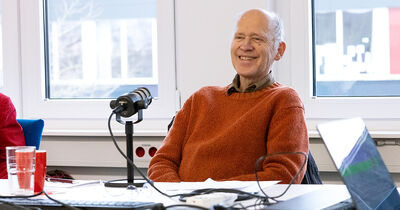 Ein Mann in einem orangenen Pullover lacht in die Kamera und hat ein Podcast-Mikro vor sich stehen. 