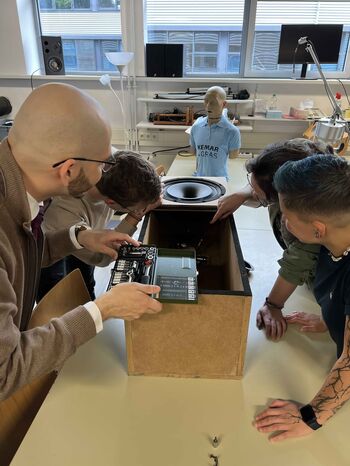 Studierendengruppe analysiert im Labor das Innenleben eines Lautsprechers mit Werkzeug, im Hintergrund ein KEMAR-Kunstkopf für Messungen.