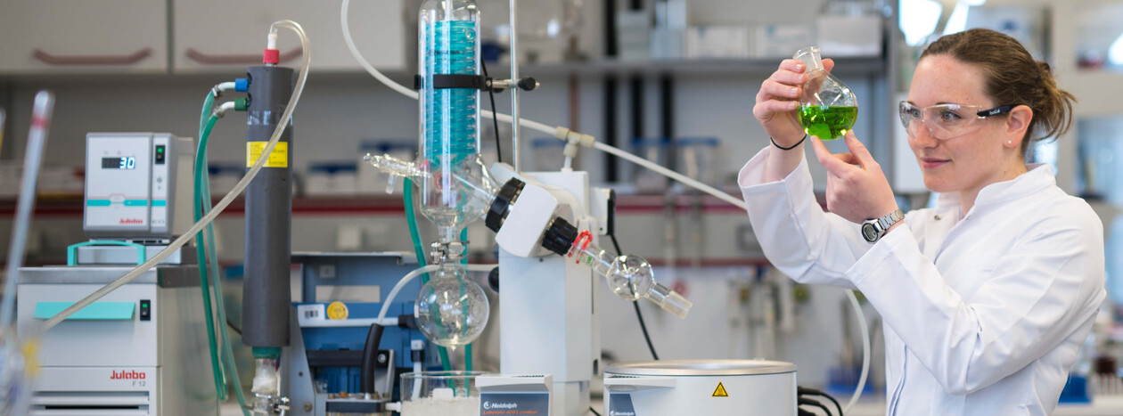 A female researcher wearing safety goggles and a lab coat examines a round-bottom flask containing a bright green liquid in a laboratory. Next to her is a complex glass apparatus.