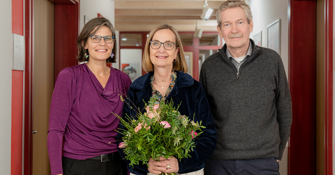 Drei Personen, links eine Frau in lila Oberteil, in der Mitte eine Frau mit Blumenstrauß und rechts ein Mann in grauem Pulli