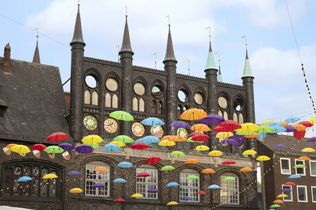 Viele bunte Regenschirme schweben vor einer historischen Backsteinfassade.