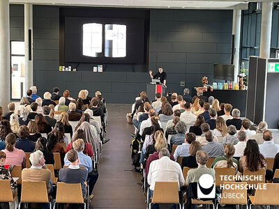 Besuchende sitzen in Stuhlreihen und gucken zur Präsentation an der Leinwand