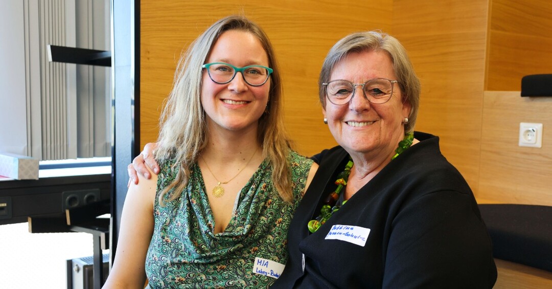 Zwei Frauen sitzen nebeneinander und lächeln in die Kamera.