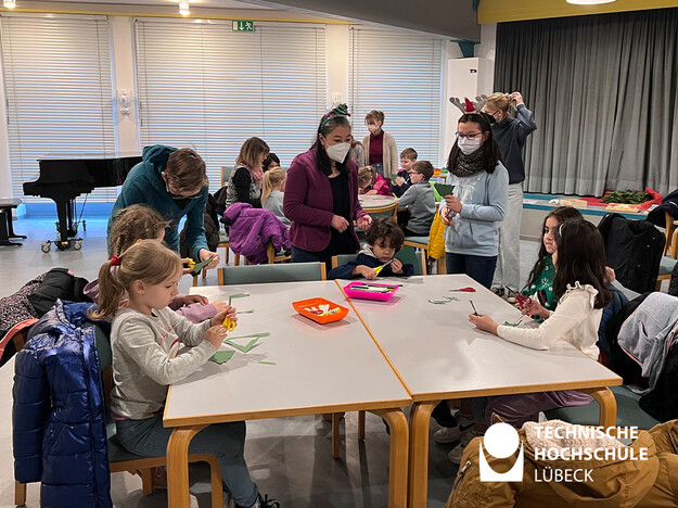 Viele Kinder sitzen an mehreren Tische und basteln eine Brille mit Hilfe von Erwachsenen