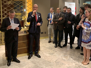Two men lead a reception; one reads from a paper while guests listen.