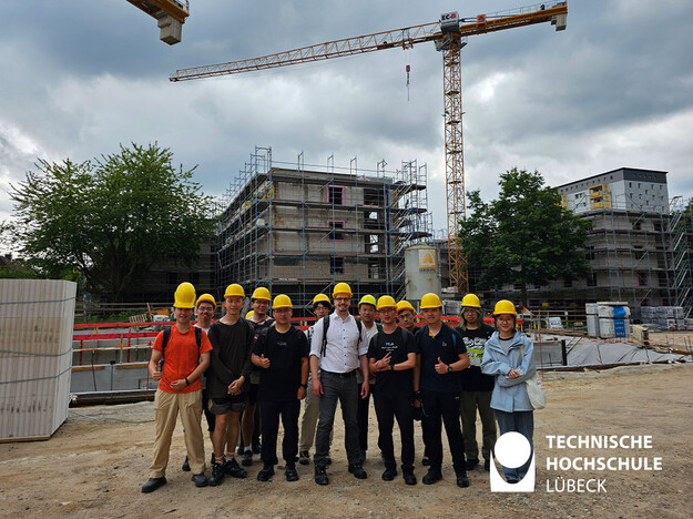 Studierende stehen auf Baustelle und im Hintergrund ein Kran und angefangenes Haus