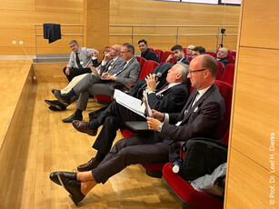 A group of men in business suits sit as an audience in a lecture hall.