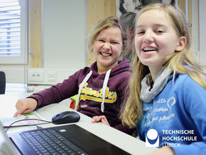 zwei Mädchen sitzen an einem Tisch vor einen Laptop und lächeln in die Kamera