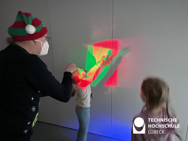 Mann leuchtet mit drei Farben auf eine Wand und Kind hält Arm in das Licht