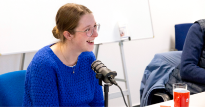 Eine Frau in blauem Pulli mit Brille sitzt vor einem Podcastmikro und lacht