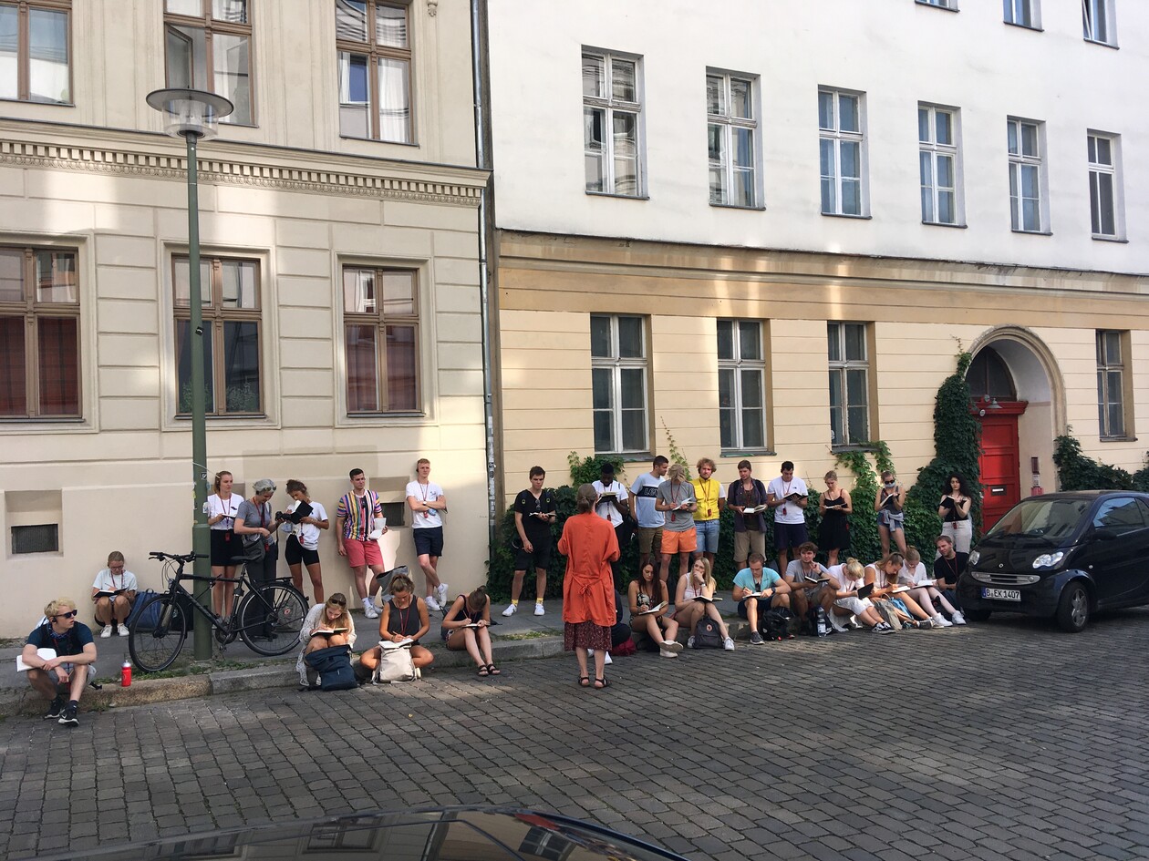 Studentengruppe zeichnet Stadtansichten auf dem Gehweg vor Altbauten.