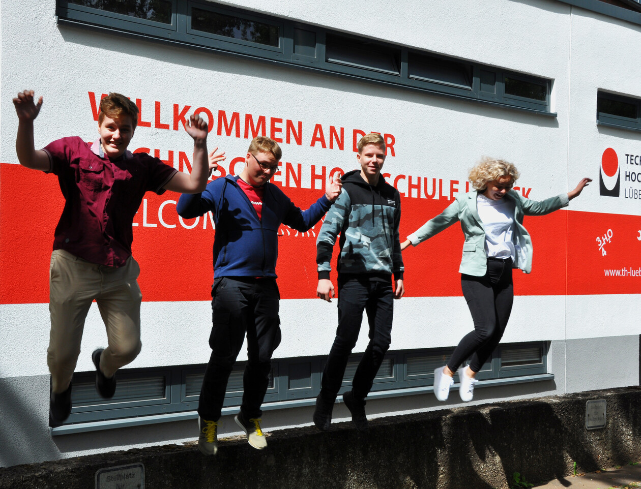 Vier junge Leute springen fröhlich vor einer Wand der TH Lübeck in die Luft.