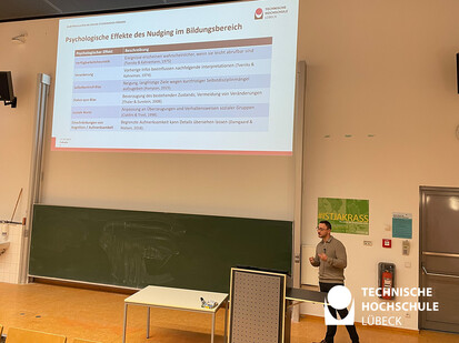 Präsentation von einem wissenschaftlichen Mitarbeiter Ein Mann steht vor einer großen Projektionsfläche und gibt eine Präsentation. Die Präsentation zeigt eine Tabelle. Der Mann steht auf der rechten Seite des Bildes.