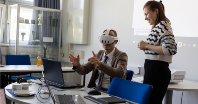 Ein Mann mit einer VR Brille greift nach etwas im Raum, eine Frau zeigt ihm wie die Anwendung funktioniert
