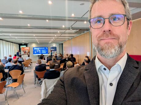 Selfie eines Teilnehmers während eines Vortrags im vollen Konferenzraum mit Präsentationsfolie im Hintergrund.