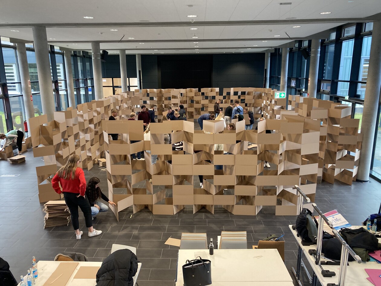 Studierende bauen eine große, begehbare Struktur aus Pappe in einer Halle.