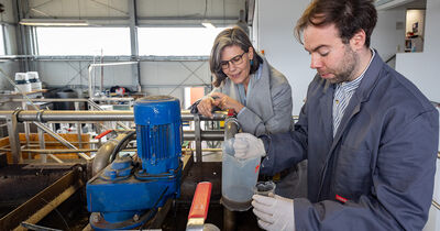 Forschungsingenieur Jascha Hödl steht zusammen mit Muriel Helbig, der Präsidentin der TH Lübeck, an der Versuchs- und Ausbildungskläranlage. Jascha Hödl füllt eine Probe des Abwassers in einen Glaszylinder.