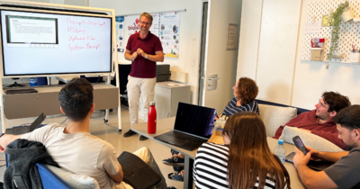Ein Professor steht vorne an einem Bildschirm, ihm schauen Studis zu
