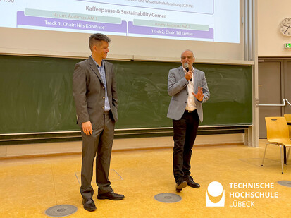 Eröffnungsrede Tag der Lehre 2023 Zwei Männer in Anzügen stehen vor einer Vorlesung. Sie sind beide in einem Bildschirm zu sehen, der auf einer Wand hängt. Der eine Mann hat eine Jacke und den anderen einen Anzug an. Sie scheinen bereit zu sein, um eine Präsentation zu halten oder zu diskutieren.
