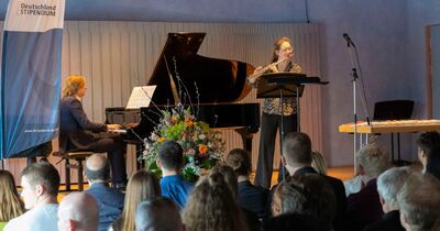 Auf dem Bild sind zwei Musiker zu sehen. Ein Mann spielt an einem Klavier und eine Frau Flöte.