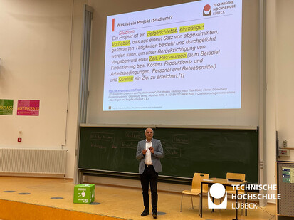 Projektmanagement und Studienerfolg Ein Mann steht vor einer großen Projektionsfläche und hält eine Präsentation. Er trägt eine graue Jacke. Die Präsentation zeigt Text auf Deutsch und ist auf einer großen Leinwand sichtbar.