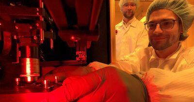 Die Doktoranden Gregor Knoll und Fabian Henk stehen am 3D-Drucker in der ein Millimeter-großes optisches Element entsteht. 