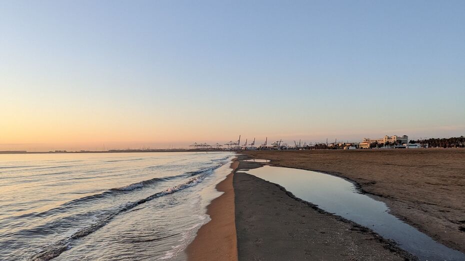 Der Sandstrand von Valencia am Abend. Der Horizont teilt das Bild mittig, auf dem in der Ferne ein Containerhafen zu sehen ist.