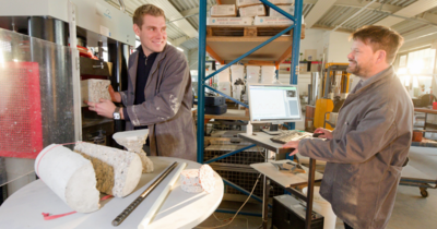 Auf dem Bild sieht man zwei Männer in grauen Kitteln, der linke steht an einer Beton Prüfmaschine und stellt einen Betonklotz hinein