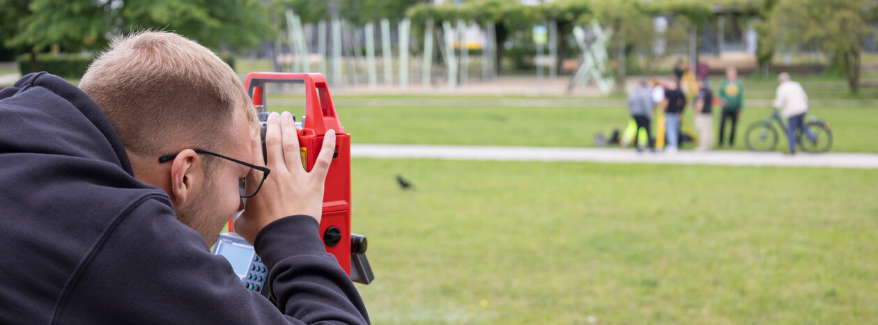 A male student, seen from the side, looks through the eyepiece of a red surveying instrument (theodolite) mounted on a tripod on a grassy area in a park.