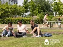 TH Lübeck Studierende und internationale Studierende sitzen auf einer Wiese und tauschen sich aus. Im Hintergrund wird Tischtennis gespielt. 