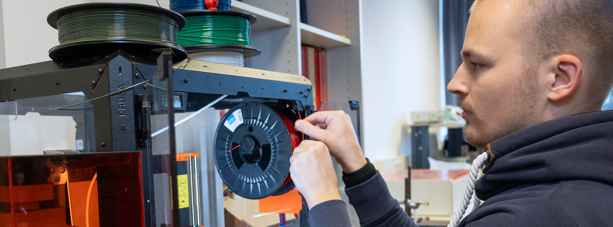 A young male student carefully threads red filament from a spool into a professional 3D printer, preparing it for a printing job.