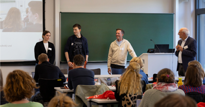 4 Personen stehen vor der Tafel und sprechen zu den Gästen
