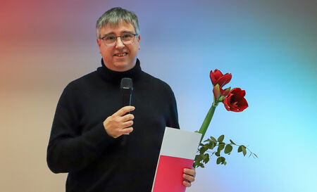 Mann mit Mikrofon und Mappe steht lächelnd mit einer roten Amaryllis.