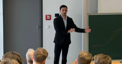 Vortragender im Anzug gestikuliert vor einer Tafel vor Publikum.