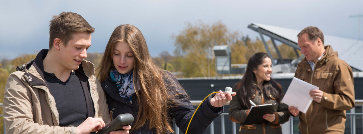 A group of students and an instructor are conducting measurements on a rooftop. A female student holds a sensor while a discussion takes place in front of a solar panel in the background.