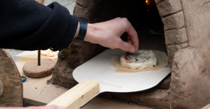 Selbstgebaute Lehmofen-Pizza auf Holzschieber, Hand schiebt Teig in den Ofen