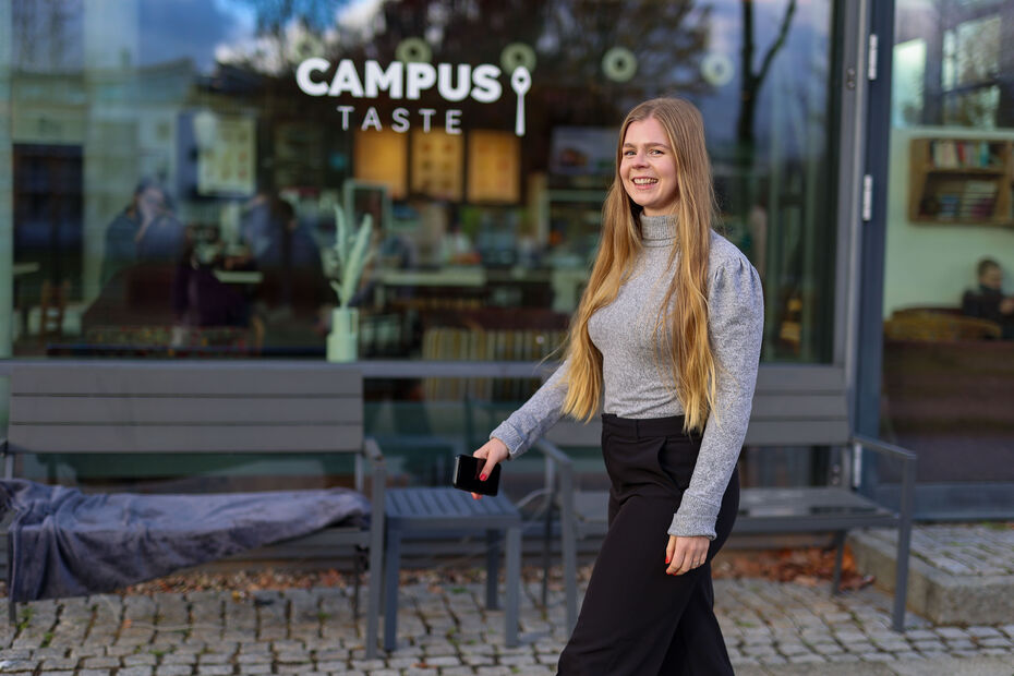 Lächelnde junge Frau steht vor der Glasfront des Cafés Campus Taste.