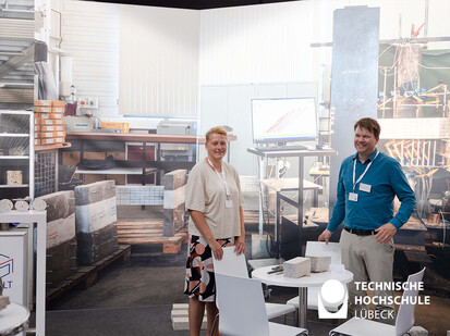Ausstellung der Materialprüfanstalt Schleswig Holstein Zwei Personen stehen an einem Tisch und im Hintergrund ein großes Plakat des Arbeitsplatzes