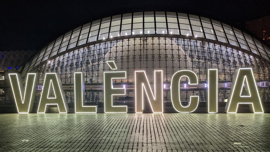 Ein Foto von einem Valencie Schriftzug, der leuchtet bei Nacht. Im Hintergrund eine erleuchtete Kuppel aus Glas