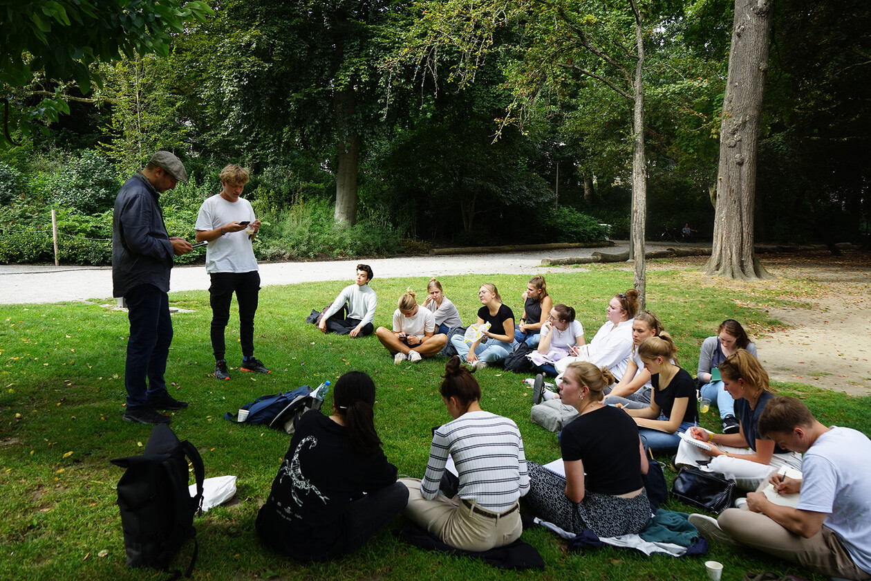 Studierende sitzen zur Besprechung im Kreis auf einer Wiese im Park.