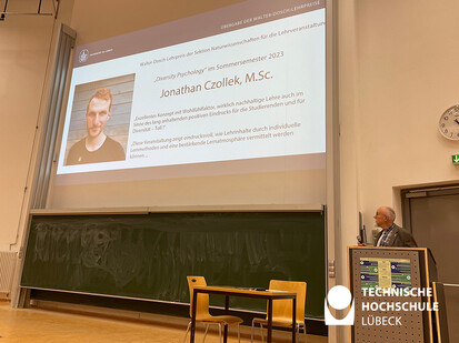 Preisübergabe der Sektion Naturwissenschaften Ein Mann in grauer Jacke steht vor einer großen Projektionsfläche und zeigt auf eine Präsentation. Die Projektionsfläche ist auf der linken Seite des Bildes zu sehen. Der Mann ist auf der rechten Seite des Bildes.