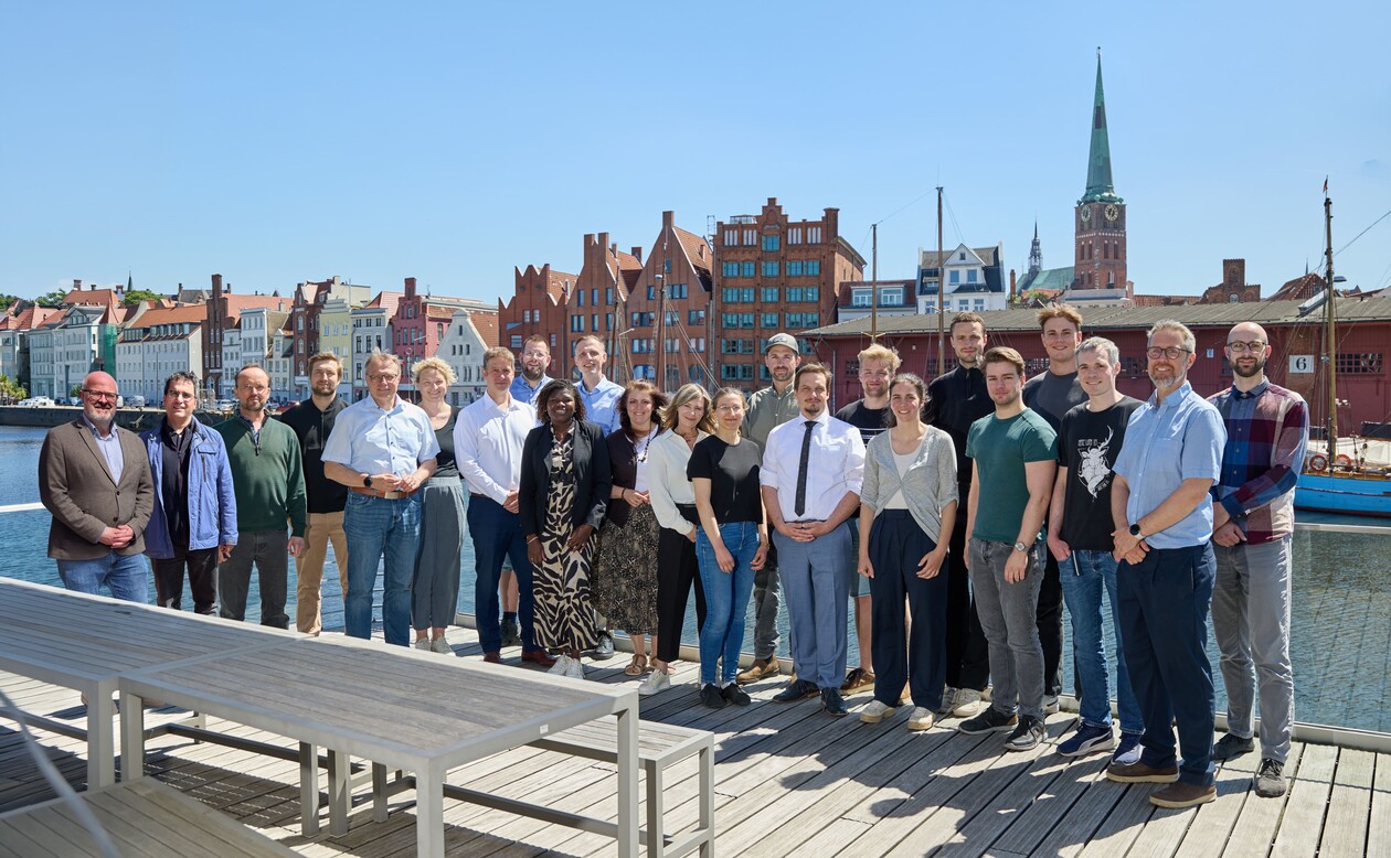 Ein Team posiert auf einer Holzterrasse am Hafen vor einer Altstadt.