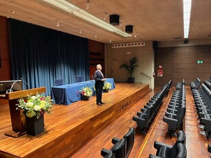 A man in a suit stands on the stage of an empty lecture hall.