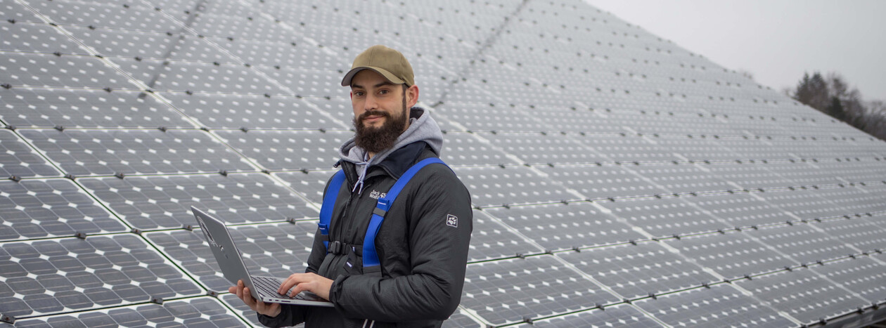 A young male student with a beard and cap holds a laptop while standing in front of a large array of solar panels, looking confidently at the camera.