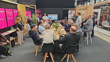 Mehrere Personen sitzen und stehen um einen großen Tisch bei einer Messe oder Tagung.