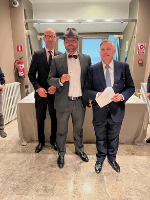 A man in a fedora and bow tie holds an award, standing with two other men.