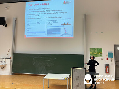 Vorstellung STUDYCoach Eine Frau steht vor einer großen Projektionsfläche und zeigt auf eine Präsentation. Die Projektionsfläche ist auf der linken Seite des Bildes zu sehen. Die Frau ist auf der rechten Seite des Bildes und hat stemmt eine Hand in die Hüfte.